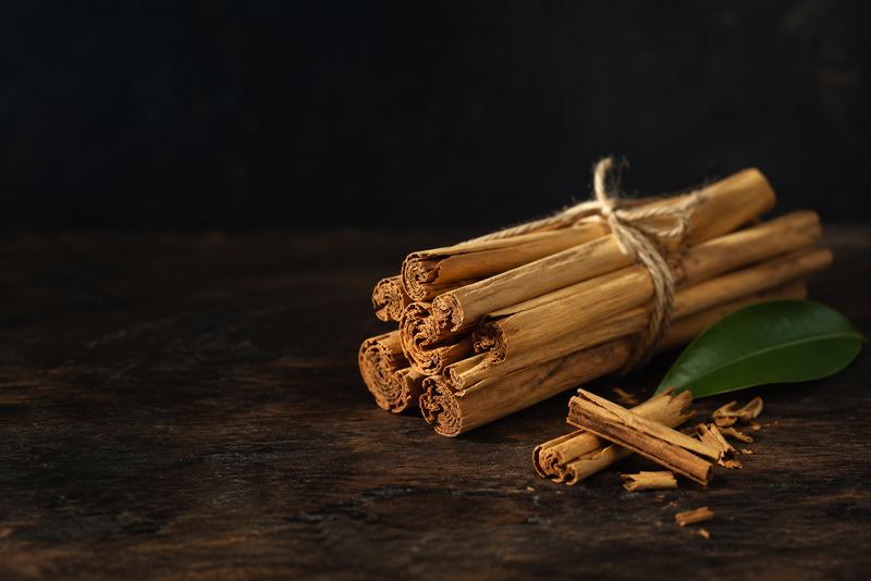 A small bundle of TRUE CEYLON CINNAMON quills tied with twine: long, thin (about a finger wide), PALE golden tan, each quill made of many paper-thin layers rolled inside each other like a fine cigar, with soft crumbly ends; NOT thick dark single-layer cassia sticks. Beside it a few loose broken quill pieces and one fresh green cinnamon leaf, on dark wood